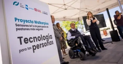 Presentación del proyecto WalkerPisa de Fundación ONCE en un paso de peatones de Las Rozas. En primer plano, un cartel con el lema “Proyecto Walkerpisa. Sensores IoT e IA para pasos de peatones más seguros. Tecnología para las personas”. Al fondo, varias personas durante el acto de presentación, una de ellas utilizando una silla de ruedas, en un entorno urbano al aire libre.