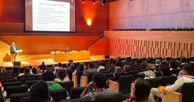 Auditorio moderno con varias decenas de personas sentadas en butacas, asistiendo a una conferencia. En el escenario, una persona realiza una presentación desde un atril mientras otra permanece sentada en una mesa de ponentes. Al fondo se proyecta una diapositiva titulada “Consecuencias del delirium”. La sala cuenta con iluminación cálida, paredes de madera y una amplia zona de gradas ocupada por el público.