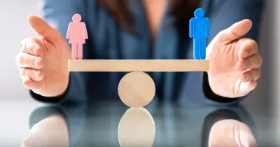 Manos sosteniendo una balanza de madera equilibrada, con una figura rosa que representa a una mujer y una figura azul que representa a un hombre, simbolizando la igualdad de género. La persona viste una blusa azul y está frente a una superficie reflectante