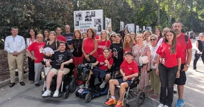 Grupo de personas reunidas al aire libre en un parque junto a varios paneles expositivos de la campaña "Escuchar cambia la historia". Algunas personas utilizan sillas de ruedas manuales y motorizadas. La imagen muestra a participantes, familiares y personas colaboradoras posando frente a la exposición fotográfica dedicada a visibilizar la realidad de quienes conviven con la distrofia muscular de Duchenne.