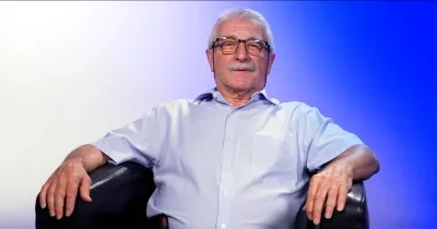 Persona sentada en un sillón negro, con gafas y camisa azul claro de manga corta, mirando al frente. El fondo presenta un degradado en tonos blancos y azules con iluminación suave de estudio. La persona aparece centrada en la imagen, con ambos brazos apoyados en los reposabrazos del sillón.