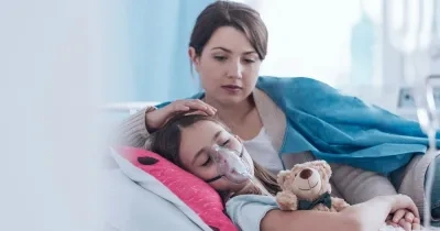 niña con una mascarilla tumbada en la cama al lado de su madre mientras la niña sufre fibrosis quítica