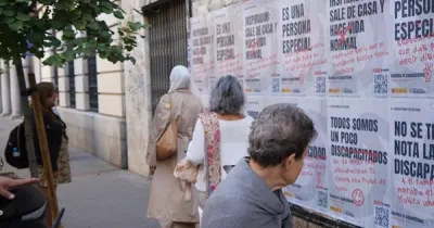 Varias personas caminan y observan una pared cubierta con carteles de una campaña contra el capacitismo en una calle de Madrid. Los carteles muestran frases asociadas a estereotipos sobre la discapacidad, algunas tachadas en rojo, junto a códigos QR y mensajes de sensibilización. En primer plano, una persona se detiene a leer los carteles, mientras al fondo se ve una hilera de árboles y edificios urbanos.