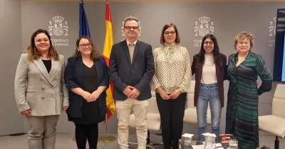 Varias personas posan de pie en una sala institucional frente a un fondo gris con el logotipo del Gobierno de España y dos banderas, una de la Unión Europea y otra de España. Delante hay una mesa con documentos, micrófonos y vasos de agua. El grupo está alineado en el centro del espacio, con sillas blancas en los laterales.