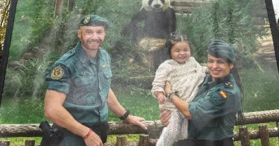 Dos miembros de la Guardia Civil posan junto a una niña delante del recinto de un panda gigante en un parque zoológico. Las tres personas se encuentran junto a una valla de madera, mientras el animal aparece al fondo rodeado de vegetación. La imagen forma parte de una acción de sensibilización vinculada al calendario solidario de la Guardia Civil.
