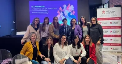 Un grupo numeroso de personas posa en una sala junto a una pantalla que muestra el título “IX Jornada Mujeres en el Autismo. Barreras y buenas prácticas”. La pantalla tiene ilustraciones de varias figuras humanas diversas. A un lado de la imagen hay un panel vertical con el logo de Federación Autismo Madrid repetido. El grupo aparece distribuido en dos filas, algunas personas de pie y otras sentadas, en un espacio interior con sillas transparentes y una mesa baja.