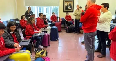 Grupo de jóvenes con sudaderas rojas sentados en un aula o sala de reuniones junto a varias maletas de viaje de distintos colores. En la parte frontal de la sala, varias personas ofrecen indicaciones antes de una salida o actividad organizada. El espacio cuenta con sillas, pupitres, grandes ventanales y equipaje distribuido por la estancia, en un ambiente de preparación para un desplazamiento colectivo.