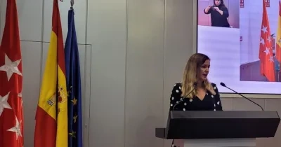 Ana Dávila, consejera de Familia, Juventud y Asuntos Sociales de la Comunidad de Madrid, subida a un atril dando un discurso con las banderas de la Comunidad de Madrid, de España y de la Unión Europea de fondo.