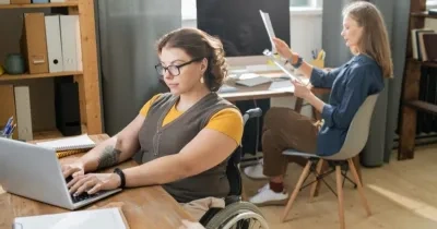 dos mujeres trabajando en sus mesas de escritorio, una de ellas tiene discapacidad física y está en silla de ruedas