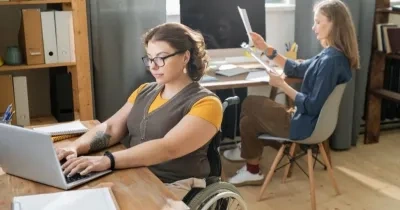 una persona con discapacidad en silla de ruedas y otra compañera sin discapacidad aparente trabajando cada una en su escritorio con su ordenador