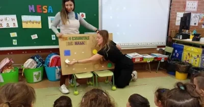 Escena en un aula infantil con dos maestras y un grupo de niños sentados en el suelo. Una maestra, vestida con una blusa gris y jeans, está de pie, mientras la otra, con ropa negra, interactúa con un modelo grande de mesa de madera con taburetes y un cartel que dice 'POR 4 ESQUINITAS DE NADA'. Fondo con pizarrón verde, dibujos, letras y materiales escolares. Ambiente colorido y educativo