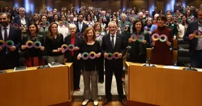 salón de actos del Congreso de los Diputados, lleno de asistentes situados tanto de pie como sentados. Todas las personas sostienen carteles con el símbolo del infinito multicolor, representativo de la diversidad del espectro del autismo. En primera fila, varios participantes permanecen de pie junto a los escaños, mientras el resto del público ocupa el fondo del auditorio. El ambiente es institucional y de apoyo colectivo a la causa del autismo.