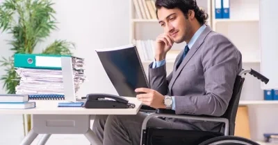 Persona con discapacidad física que va en silla de ruedas traajando en el escritorio con su ordenador y cuadernos.