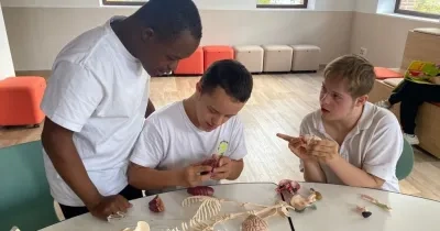 dos niños con síndrome de down junto a su profesor mientras colocan las piezas de un esqueleto de juguete para saber las partes del cuerpo humano