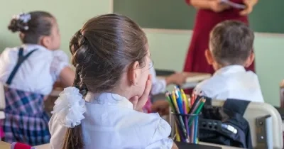 tres niños en una escuela sentados en un pupitre de espaldas a la imagen y de fondo la profesora dando clase.