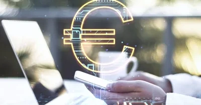 Mano sosteniendo un teléfono móvil junto a un ordenador portátil, con un símbolo del euro digital superpuesto en el centro de la imagen. La escena representa conceptos relacionados con pagos electrónicos, finanzas digitales y tecnología financiera. Fondo desenfocado con tonos claros y elementos tecnológicos.