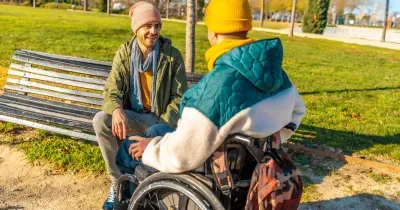 Persona en silla de ruedas y otra persona mantienen una conversación en un parque urbano con zonas verdes y caminos peatonales.