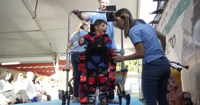 Niño utilizando un exoesqueleto pediátrico asistido por profesionales durante una demostración de neurorrehabilitación. El menor se encuentra sujeto a un dispositivo robótico de marcha sobre una plataforma, mientras personal especializado supervisa y ajusta el equipo ante un público asistente.