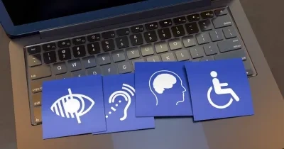 Cuatro post-its azules con símbolos de accesibilidad colocados sobre el teclado de una laptop gris. Los símbolos representan: un ojo con líneas (visión), una oreja con ondas (audición), un perfil de cabeza (cognición) y una persona en una silla de ruedas (movilidad). La pantalla de la laptop está en negro