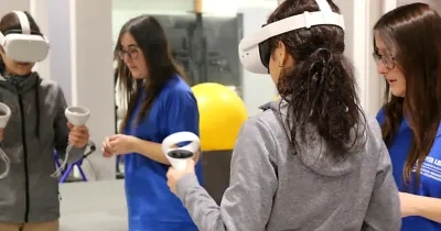 Persona utilizando unas gafas de realidad virtual y mandos de control durante una actividad de rehabilitación tecnológica en un centro especializado. En la sala hay otras dos profesionales con uniforme azul y una pelota terapéutica amarilla al fondo.