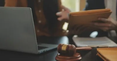 delante de la imagen podemos ver un mazo de juez y un ordenador portátil, al fondo observamos a dos personas haciendo un intercambio de una carpeta de documentos
