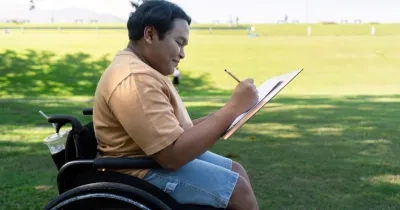 Niño en silla de ruedas escribiendo en un cuaderno a la sombra en un césped