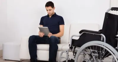 chico con discapacidad sentado en el sofa usando una tablet, al lado suya está su silla de ruedas
