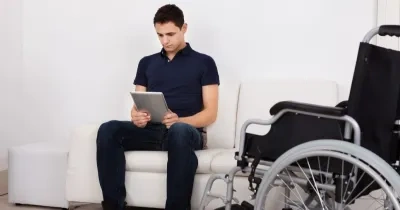 Persona con movilidad reducida sentado en el sillón junto a su silla de ruedas, mientras usa la tablet