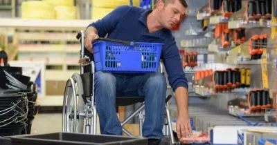 Interior de una tienda de bricolaje o ferretería. Una persona en silla de ruedas se desplaza por un pasillo comercial mientras sostiene una cesta de compra azul sobre las piernas. Con una mano alcanza un producto situado en un estante inferior. A ambos lados del pasillo hay estanterías con herramientas, accesorios y materiales organizados. La imagen muestra una situación de compra y accesibilidad en un entorno comercial.