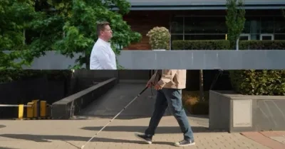 Dos personas aparecen en un entorno al aire libre con un edificio moderno y vegetación de fondo. La persona de la izquierda, vestida con una camisa blanca, está de pie cerca de un seto y árboles. La persona de la derecha, con una chaqueta beige, jeans azules y zapatillas, camina con un bastón blanco, navegando por un sendero pavimentado con una rampa y un muro bajo
