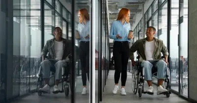 Chico con silla de ruedas hablando por la oficina con una compañera sin aparente discapacidad