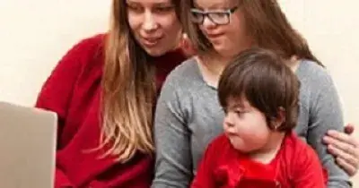 Una persona enseñando a manejar un ordenador a otras dos con discapacidad cognitiva (una adolescente y un bebé)