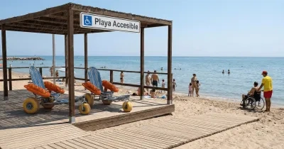 **Alt text:**  Playa con servicio accesible señalizado (“Playa Accesible”) bajo una estructura de madera con pasarela hasta la orilla, donde hay varias sillas anfibias con flotadores naranjas; al fondo, personas en la arena y baño en el mar, y a la derecha una persona usuaria de silla de ruedas acompañada junto al agua.