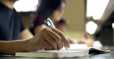 un chico escribiendo en una libreta mientras de fondo salen dos personas estudiando en la biblioteca