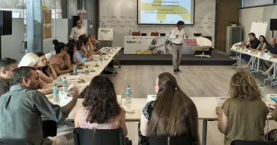 Muchas personas sentadas en una mesa con una ponente en el medio que modera y dirige la sesión