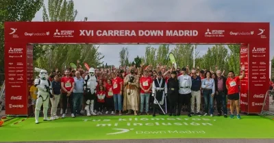 Participantes de la XVI Carrera Down Madrid posan bajo el arco de salida en el Parque Juan Carlos I antes del inicio de la prueba. En primer plano aparecen corredores, voluntarios y personas disfrazadas de personajes de ciencia ficción, mientras una gran multitud se concentra detrás. El arco rojo muestra el nombre del evento y los logotipos de las entidades colaboradoras. La carrera tiene como objetivo promover la inclusión de las personas con discapacidad intelectual a través del deporte.