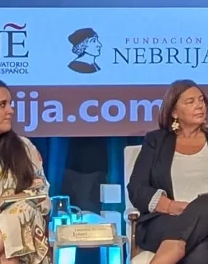 Público asistente y participantes en la presentación del libro Lectura Fácil y web comprensible. Un libro blanco en el marco del nuevo reglamento, celebrada en el Salón de Grados del Campus de Princesa de la Universidad Nebrija. En el escenario, varias personas intervienen en una mesa redonda mientras una persona realiza una presentación desde un atril. Al fondo se proyecta la imagen del evento y se muestran banderas institucionales.