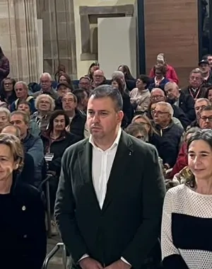 Salón de actos instalado en el interior de un edificio histórico con altos arcos y columnas de piedra. En primer plano, seis personas permanecen de pie frente a una mesa con micrófonos, documentos y botellas de agua. Detrás, decenas de asistentes ocupan las filas de asientos durante un acto público. La estancia está iluminada por luz artificial y por un gran ventanal circular situado en la parte superior del fondo de la sala. El público y las personas situadas en la mesa se distribuyen bajo una arquitectura