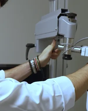 Profesional sanitario realiza una prueba de visión a una persona sentada utilizando un equipo optométrico en una consulta. La persona examinada lleva colocado un dispositivo frente a los ojos mientras el especialista ajusta el aparato para evaluar su capacidad visual. El entorno es una sala clínica con equipamiento oftalmológico.
