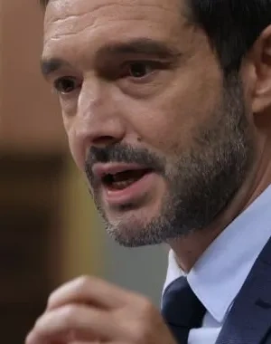 Persona con traje oscuro interviene desde un atril con dos micrófonos en el interior de una cámara parlamentaria. La imagen muestra a la persona realizando un gesto con la mano mientras se dirige al público, con el fondo desenfocado y elementos arquitectónicos de la sala visibles.