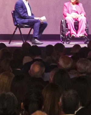 Dos personas subidas en el escenario hablando sentados en dos sillas, mientras hay multitud de personas en la grada observando 