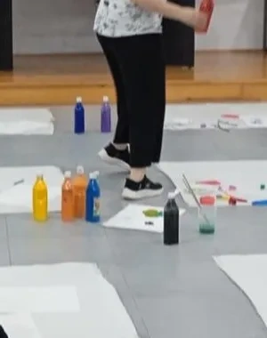Alt text:  Grupo de menores participa en una actividad artística inclusiva en una sala escolar, pintando sobre grandes hojas de papel colocadas en el suelo. Varias personas trabajan con pinceles y pintura de colores mientras una profesional supervisa la actividad. La imagen refleja un entorno educativo colaborativo orientado al aprendizaje y la participación.