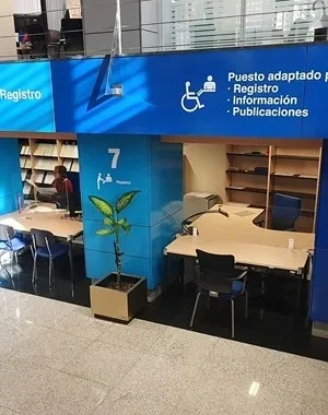 Vista interior de la Oficina de Información y Registro del Paseo del Prado, con varios puestos numerados (6, 7 y 8) señalizados con grandes paneles azules. Cada puesto incluye pictogramas, colores diferenciados y símbolos de accesibilidad. Se observan mesas, sillas, estanterías y un ascensor cercano. La señalética es clara y de alto contraste para facilitar la orientación.