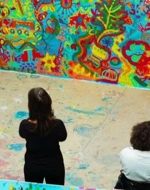 Foto Creación Ilusión con varias personas presenciando la obra, que es un mural grande con mucho colorido.