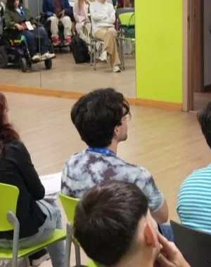 Grupo de personas asistiendo a una sesión formativa o conferencia en una sala accesible. Una persona realiza una presentación frente al público junto a una pantalla, mientras otra persona interpreta en lengua de signos. Entre los asistentes hay personas usuarias de silla de ruedas. La sala cuenta con paredes de colores vivos y un mural de cascadas al fondo.