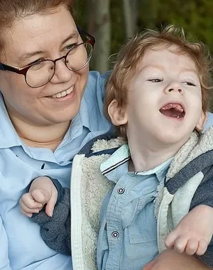 Persona adulta con gafas sostiene a un niño con discapacidad en un columpio adaptado al aire libre. Ambos se encuentran en una zona de juego rodeada de árboles. La imagen refleja acompañamiento, inclusión y apoyo a la infancia con discapacidad en un entorno accesible y seguro.