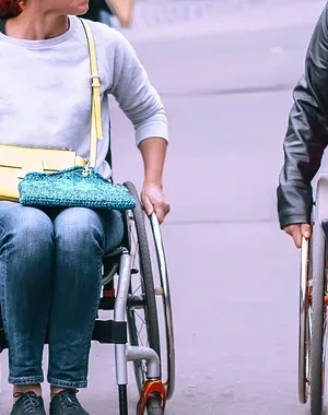 Dos personas usuarias de silla de ruedas se desplazan por una vía urbana peatonal. Una de ellas viste una camiseta clara y lleva un bolso cruzado, mientras que la otra viste un vestido rojo y una chaqueta oscura. A su alrededor caminan otras personas y se observan vehículos aparcados, árboles y mobiliario urbano al fondo. La escena transcurre al aire libre en un entorno urbano accesible.