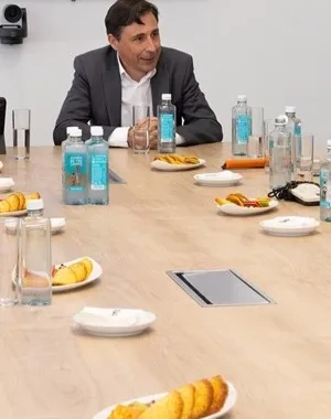 Varios representantes sentados en una mesa hablando, se observan papeles, botellas de agua y comida en el centro de la mesa
