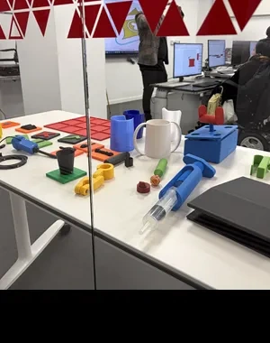 Mesa de trabajo con diversos objetos fabricados mediante impresión 3D, entre ellos recipientes, piezas de ensamblaje, una taza, adaptadores y herramientas de apoyo en distintos colores. Al fondo, tras un cristal decorado con triángulos rojos, varias personas participan en una actividad formativa frente a ordenadores mientras observan diseños digitales mostrados en las pantallas.