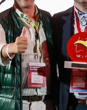 Tres personas posan sobre un escenario durante un acto de Startup Olé Salamanca. Una de ellas sostiene un premio de color rojo mientras otra hace el gesto de aprobación con el pulgar. En primer plano hay varios micrófonos y, al fondo, se aprecia una batería y el logotipo del evento proyectado en una pantalla.